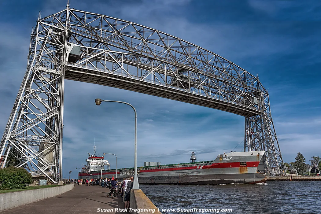   
The cargo ship Beatrix passes under the Duluth Aerial Lift Bridge as she enters the harbor. People stand along the waterfront watching the ship, and a lighthouse is visible near the end of the pier under a mostly clear sky.
