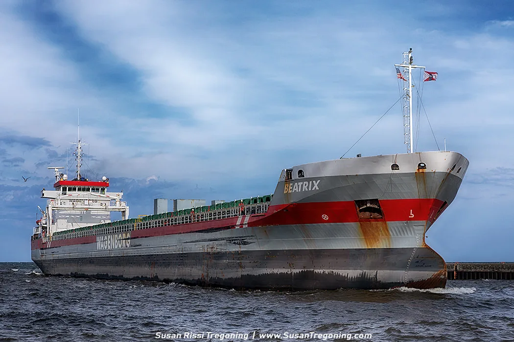 The cargo ship Beatrix enters the Duluth Ship Canal, moving between the piers under a partly cloudy sky. The ship has a gray and red hull with visible rust near the bow, and its deck cargo is covered with green tarps as birds fly nearby.