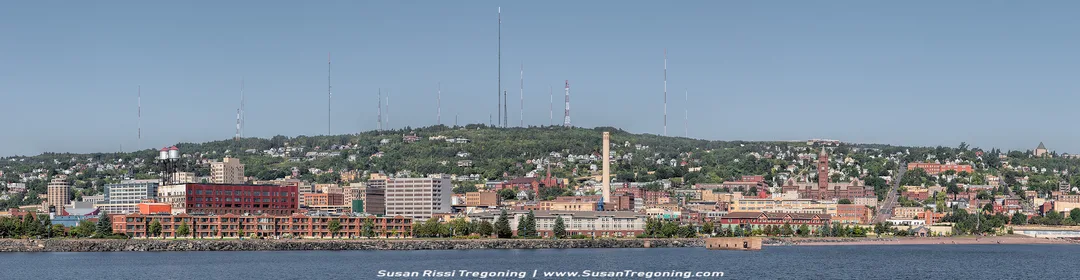 A panoramic view of Duluth, Minnesota, showing the city built along the Lake Superior waterfront with neighborhoods rising up the hillside. Numerous communication towers line the ridge above the city. The scene includes a mix of modern buildings, older brick structures, and industrial areas, with calm water in the foreground and a clear blue sky overhead.