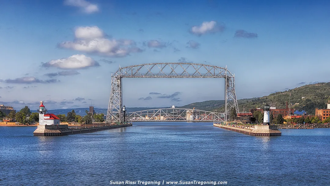 A view of the Aerial Lift Bridge in Duluth, Minnesota, seen from the water as the canal opens toward Lake Superior. The steel lift span rises above the Duluth Ship Canal, with the two pier lighthouses on either side—one with a red roof on the left and a white tower on the right. Hillside buildings and a partly cloudy sky appear in the background.
