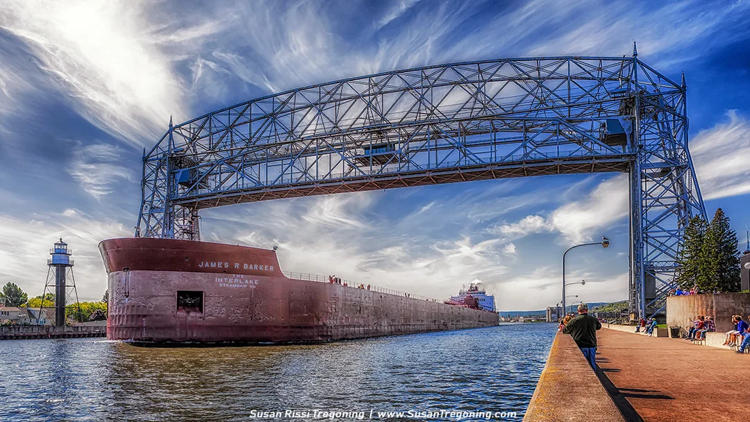 The cargo ship James R. Barker passes beneath the Aerial Lift Bridge, with a lighthouse on the left and people gathered along the canal walkway on the right. The ship fills much of the channel as it departs, and dramatic, wispy clouds stretch across the bright sky above.