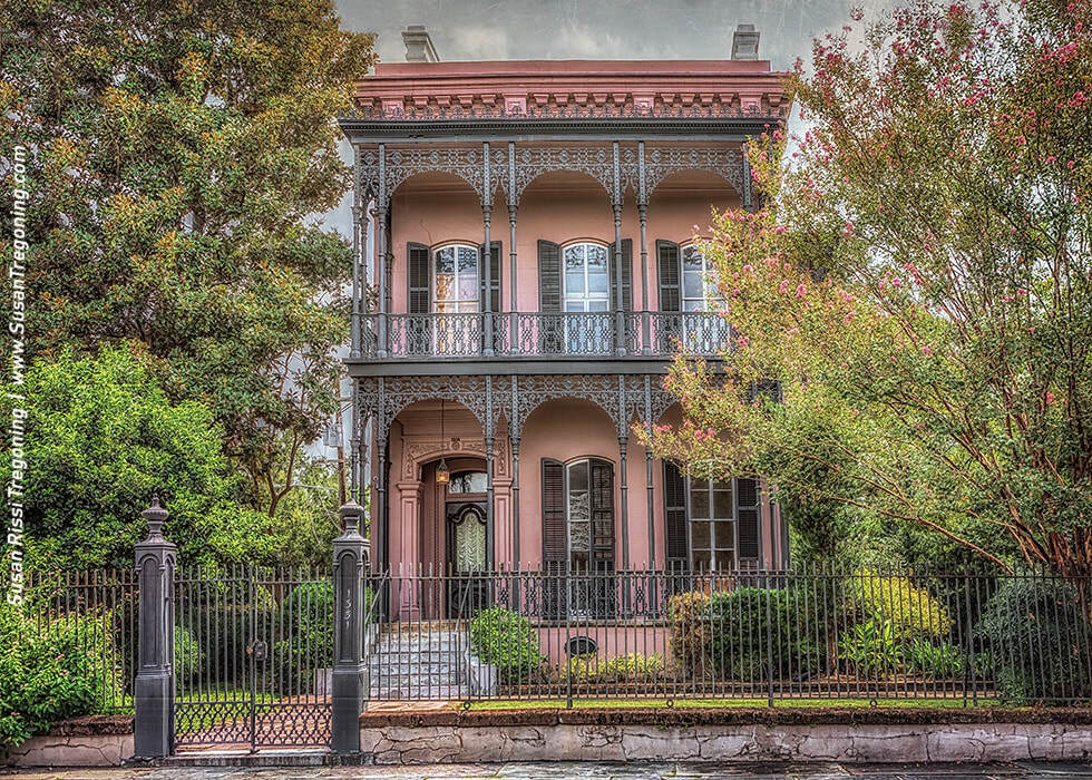 A two‑story historic house with a pink exterior features tall shuttered windows, ornate wrought‑iron balconies, and a central doorway framed by decorative ironwork. Lush greenery surrounds the home, and a black iron fence encloses the property, highlighting its symmetrical Victorian‑era design.