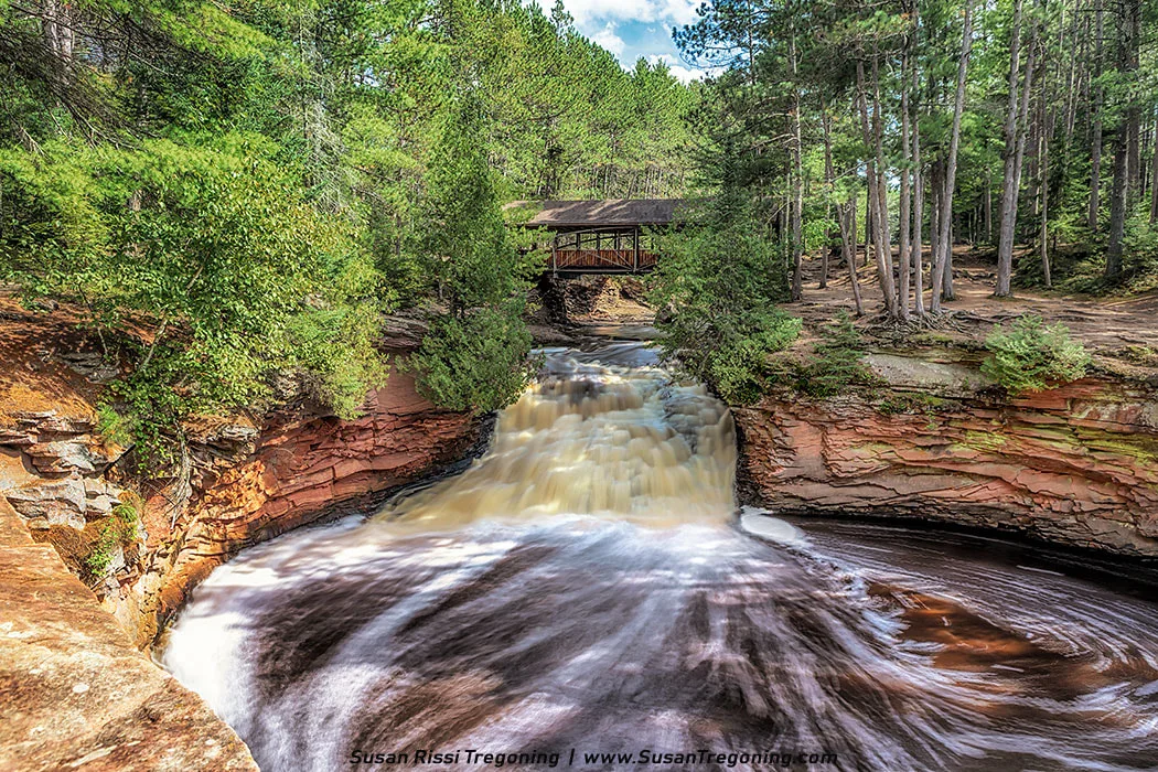 A waterfall flows over layered red rock into a swirling pool below, with a rustic covered wooden bridge spanning the stream above. Surrounded by tall green trees and forested banks, the scene captures the motion of the water with a soft, long-exposure effect under bright daylight.