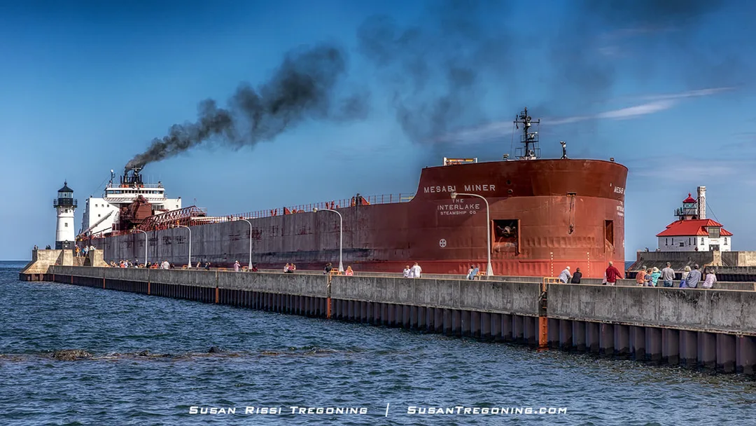    
A large Great Lakes freighter named Mesabi Miner moves through the Duluth Ship Canal on a clear day, with visitors standing along both piers and the canal’s two lighthouses visible on either side.