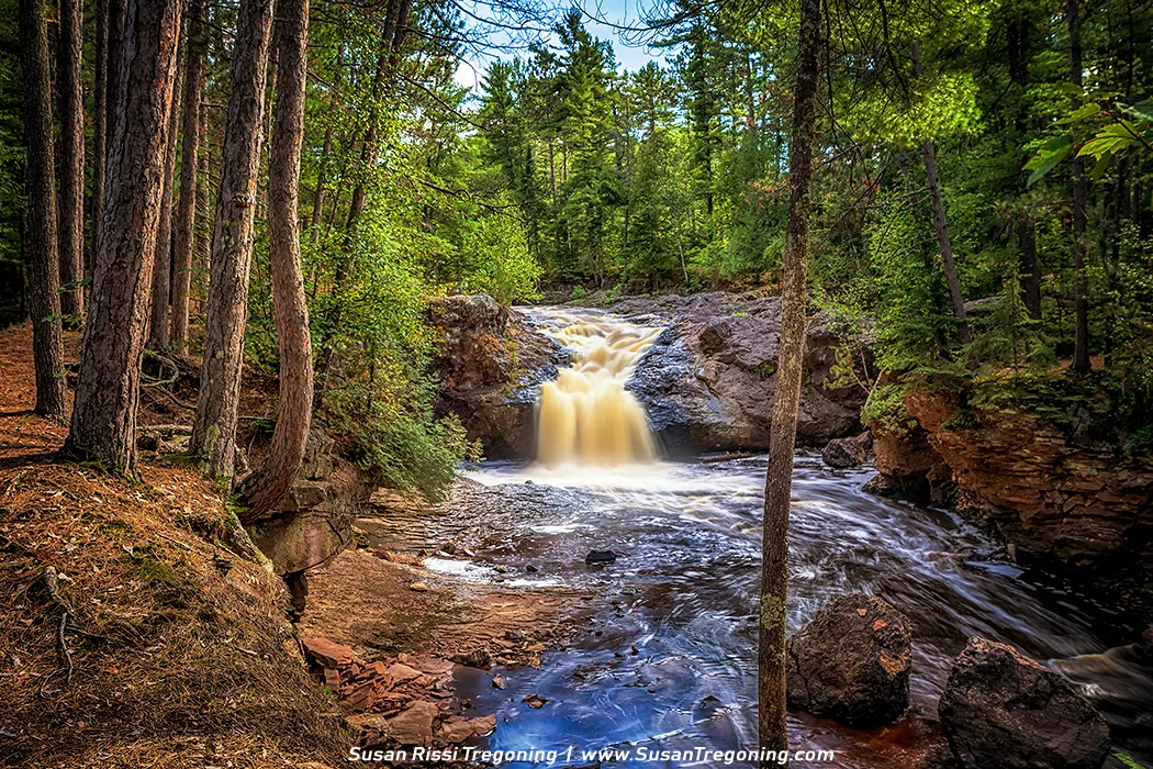 A long‑exposure view of Upper Amnicon Falls in late summer, where the Amnicon River drops over a broad rock ledge into a shallow pool. The timed exposure creates silky, blurred water surrounded by lush green forest.