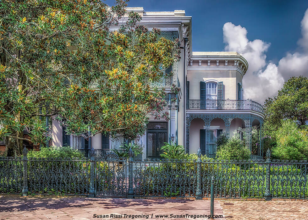 A large 19th‑century mansion with a light exterior and tall shuttered windows stands behind an ornate wrought‑iron balcony and decorative fence. Lush greenery and trees surround the home, and a brick walkway leads toward the entrance. The partly cloudy sky adds contrast above the historic Garden District residence.