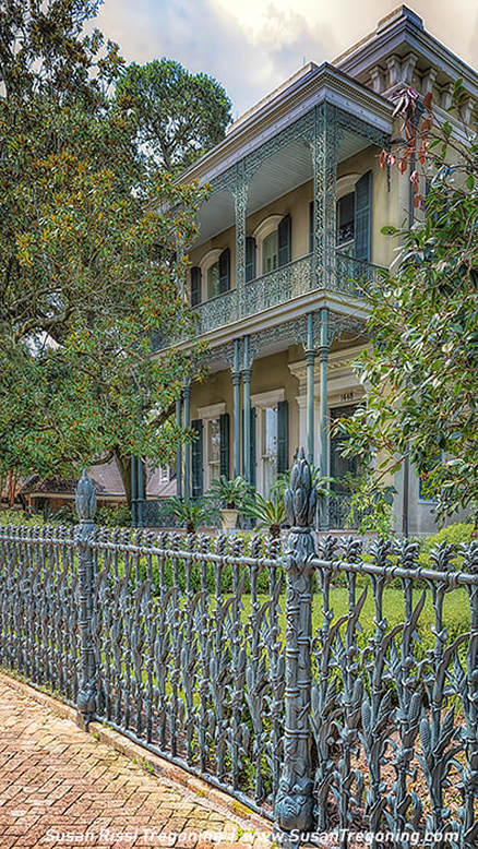A two‑story historic mansion features tall columns, shuttered windows, and ornate wrought‑iron railings on the balcony and fence. Lush greenery and trees surround the home, and a brick walkway runs along the front of the property.