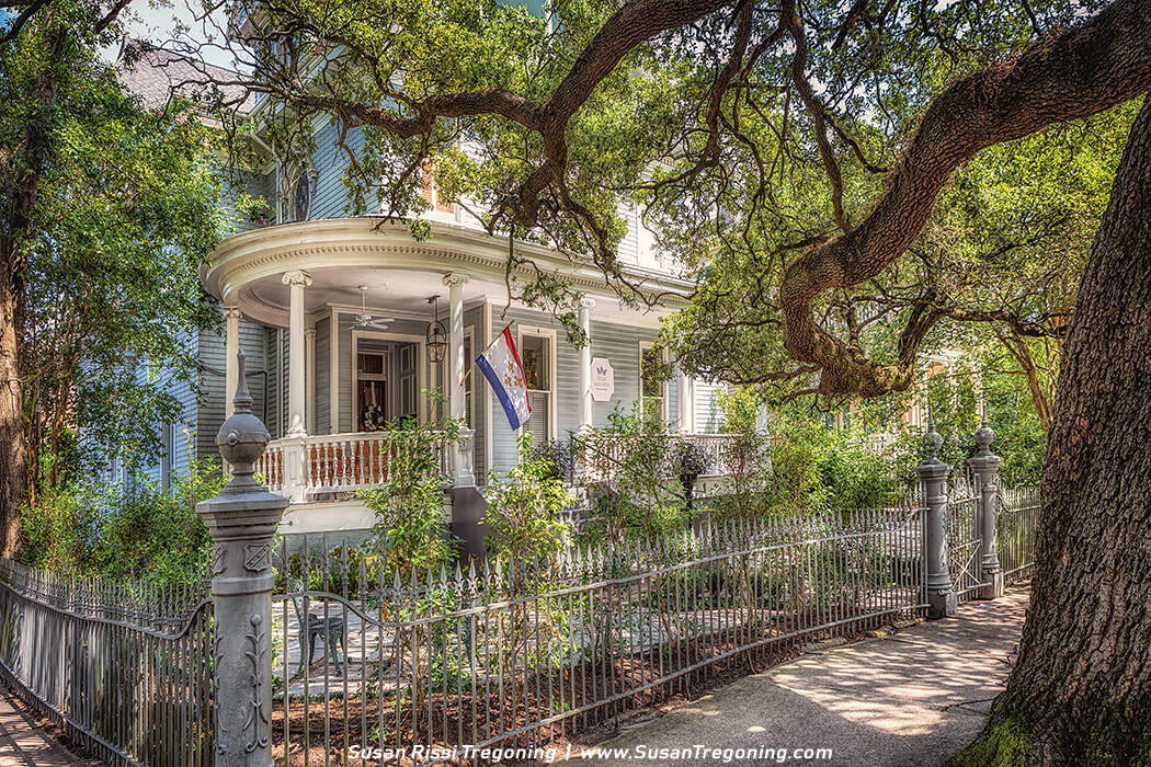A large historic house with light blue‑gray siding and white trim features a rounded front porch supported by white columns, ornate railings, and a hanging lantern. A red, white, and blue flag hangs near the entrance, and a decorative wrought‑iron fence and lush trees frame the shaded residential scene.