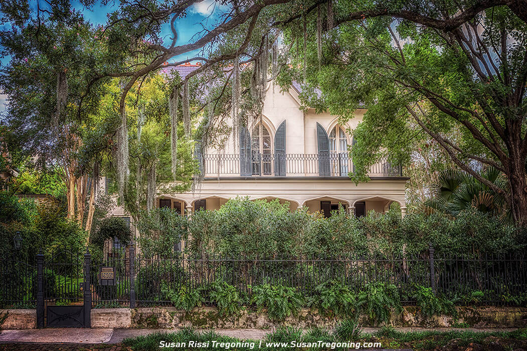 A large two‑story historic house is partially hidden by lush greenery and tall trees draped with Spanish moss. The home features tall arched windows with dark shutters, a wrought‑iron balcony railing, and a columned porch. A black iron fence and dense foliage line the front of the property.