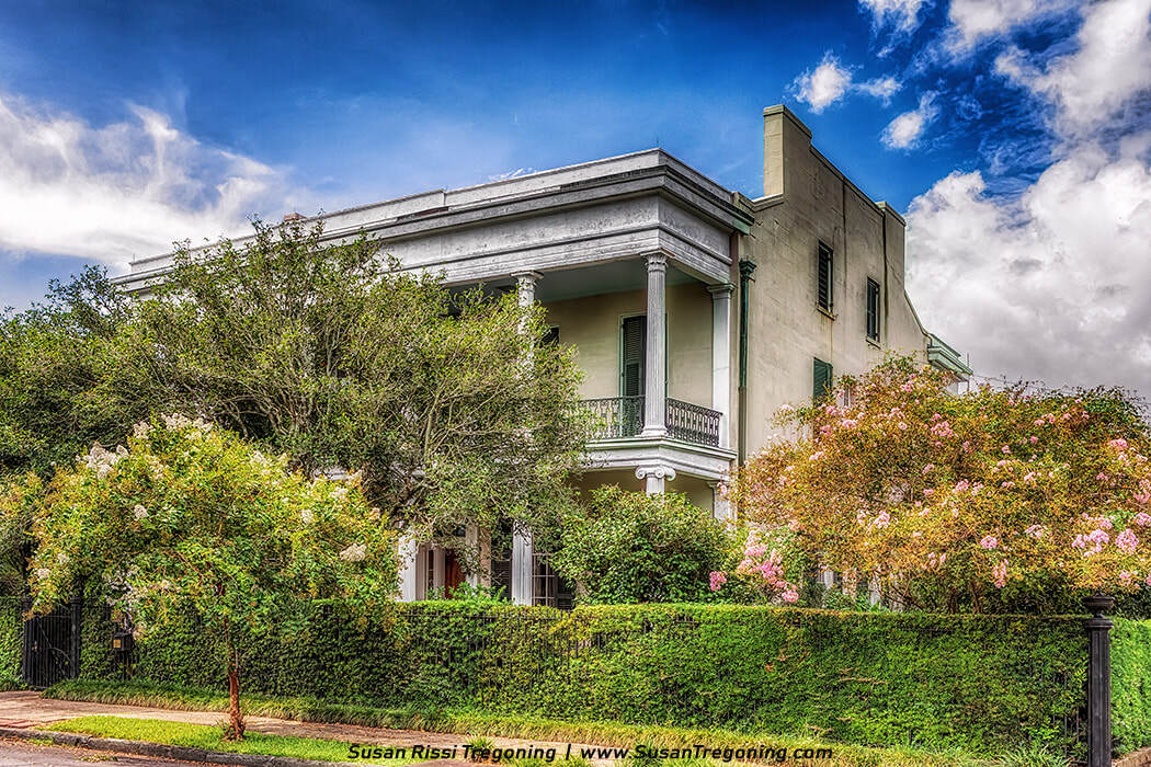 A large two‑story historic house with tall white columns and a wraparound balcony features green shuttered windows and a light‑colored exterior. Dense hedges, flowering shrubs, and mature trees surround the home, with branches framing the façade against a partly cloudy sky.