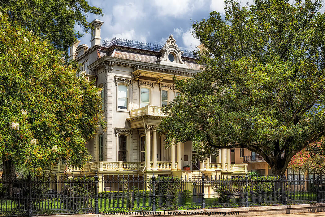 A large historic mansion painted in light cream and beige features tall classical columns, decorative cornices, intricate moldings, and iron cresting along the roofline. Mature trees and a manicured lawn surround the house, and a black wrought‑iron fence encloses the property.