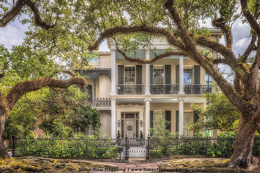 A large two‑story historic house with tall white columns and a full‑width balcony features ornate wrought‑iron railings, symmetrical shuttered windows, and a centered front door flanked by lantern lights. Mature trees with curving branches frame the scene, while a brick pathway and decorative iron fence lead to the entrance, surrounded by lush greenery.