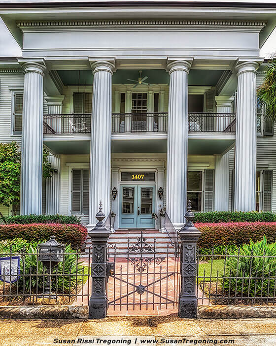 A large two‑story house with tall white columns and a wide porch features a light blue front door marked with the number 1407. Ornate wrought‑iron fencing and posts frame the entrance, with neatly trimmed shrubs and a brick walkway leading to the gate. The symmetrical façade includes shuttered windows and a balcony supported by the columns.