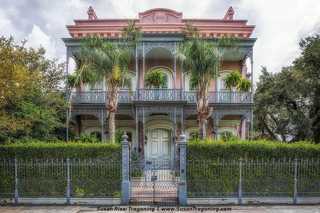 A large two‑story pink historic house features ornate wrought‑iron balconies, arched tall windows, and a central double‑door entrance framed by columns. Hanging plants decorate the upper balcony, while palm trees and manicured hedges surround the façade. A black wrought‑iron fence encloses the property, emphasizing its symmetrical Victorian‑era design.