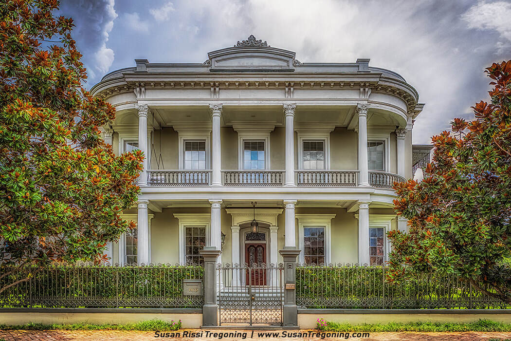 A grand two‑story mansion with a light exterior features tall white columns supporting two levels of porches, ornate railings, and a decorative pediment above the central entrance. A wrought‑iron fence encloses the property, and large magnolia trees frame the house beneath a partly cloudy sky.