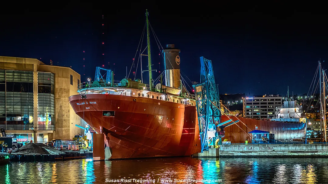 Crews move the SS William A. Irvin out of the Minnesota Slip at night, guiding the ship through the narrow Minnesota Slip Bridge using winches. The freighter is centered between the tight bridge walls, with bright work lights illuminating the ves