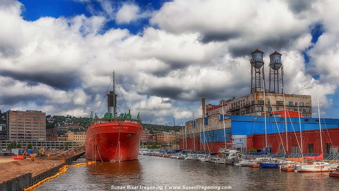 The 610‑foot Great Lakes freighter SS William A. Irvin is docked along the waterfront, with its long hull and deck structures visible beneath cloudy skies. Industrial buildings and museum‑ship infrastructure surround the vessel.