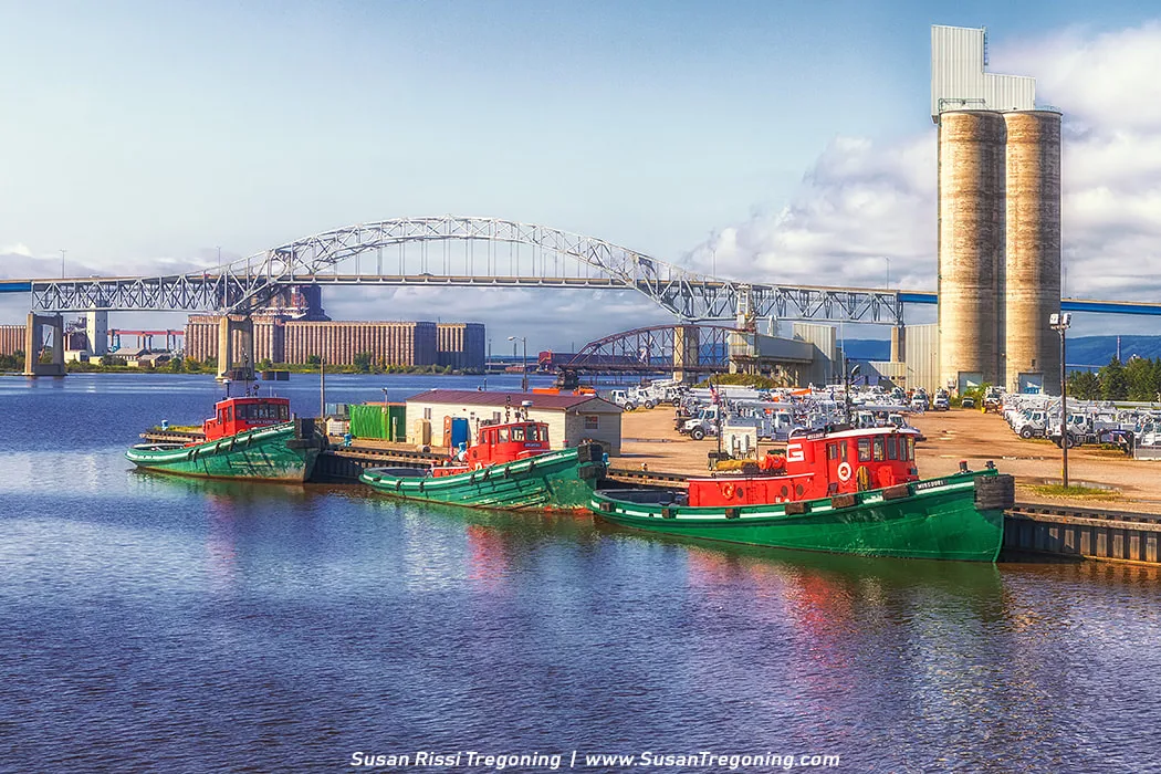   
Three green and red tugboats from the Great Lakes Towing Company are moored along a calm waterfront at Rice Point. Utility vehicles and small buildings sit on the dock behind them, and the arched steel span of the John A. Blatnik Bridge rises in the background. Farther back, tall silos and industrial structures line the harbor under a partly cloudy sky.