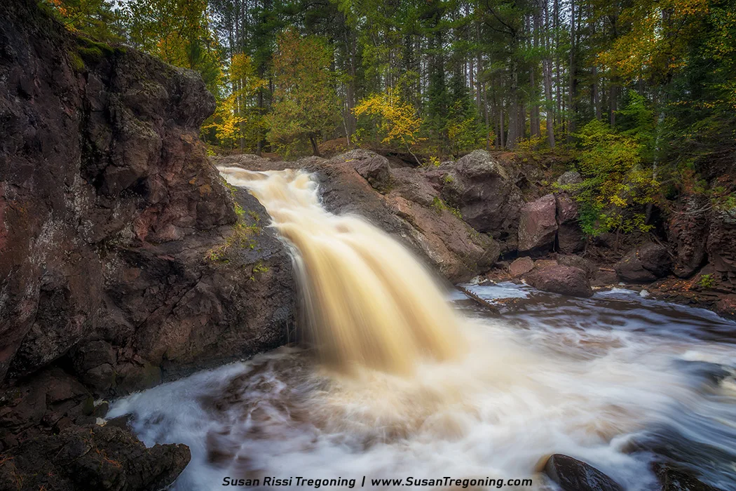 A small waterfall cascades over dark, rugged rocks into a swirling pool below, surrounded by a dense forest of tall trees with hints of early autumn foliage. The water appears smooth and misty from a long-exposure effect, and the scene has a calm, natural atmosphere.