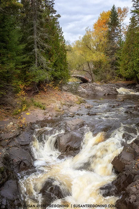 The beginning of The Deeps at Lester Park, where a small waterfall spills over dark rocks into a narrow forested gorge. Evergreen trees and early autumn foliage frame the rushing water, with a stone arch bridge partially visible through the trees under a cloudy sky.