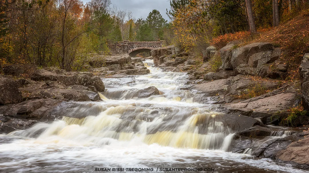 A cascading waterfall flows over dark rocks beneath Bridge #6 on Seven Bridges Road. The stone arch bridge rises above the stream, surrounded by dense autumn foliage in shades of orange, yellow, and green.