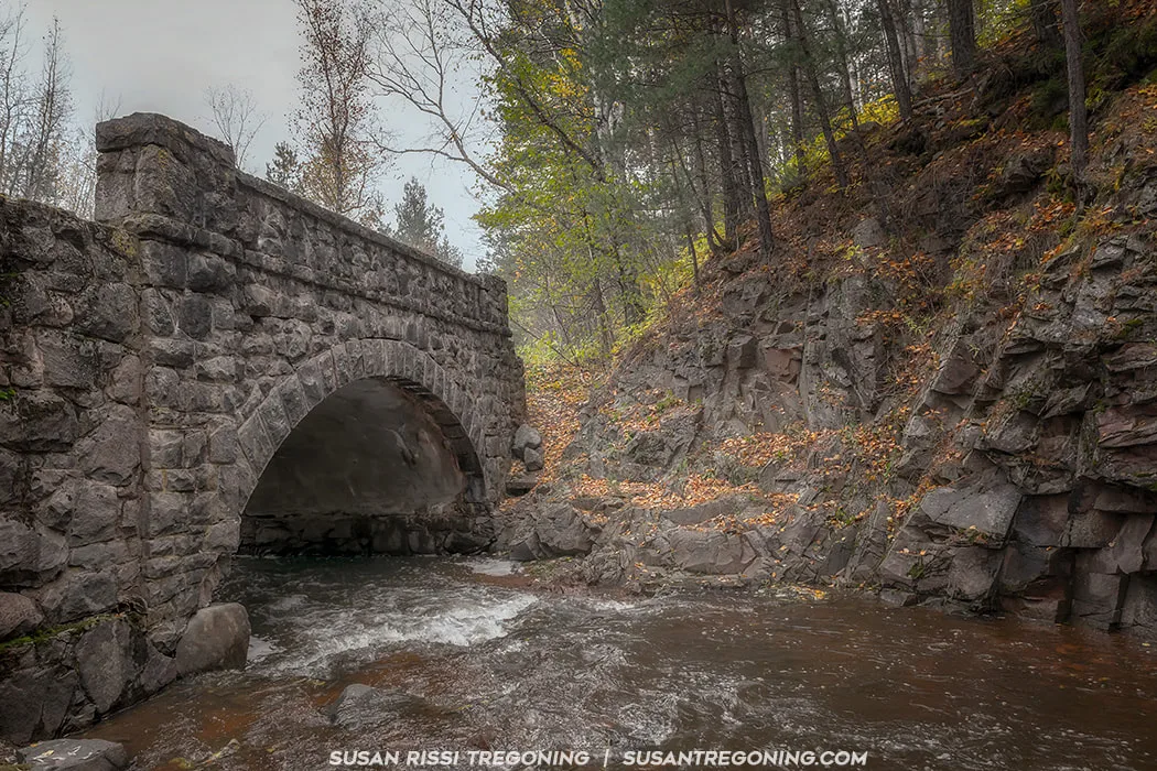 Bridge #8 on Seven Bridges Road, an original early‑1900s stone arch, spans a rocky stream in a steep forested gorge. Water rushes beneath the bridge, surrounded by autumn leaves and rugged terrain.