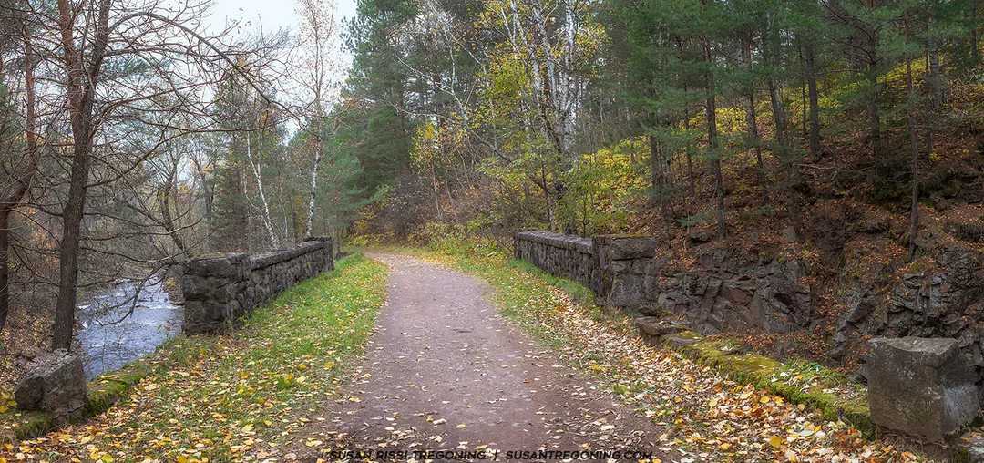 A leaf‑covered forest path crosses a small stone bridge beside a flowing creek, surrounded by late‑autumn trees and rocky terrain.