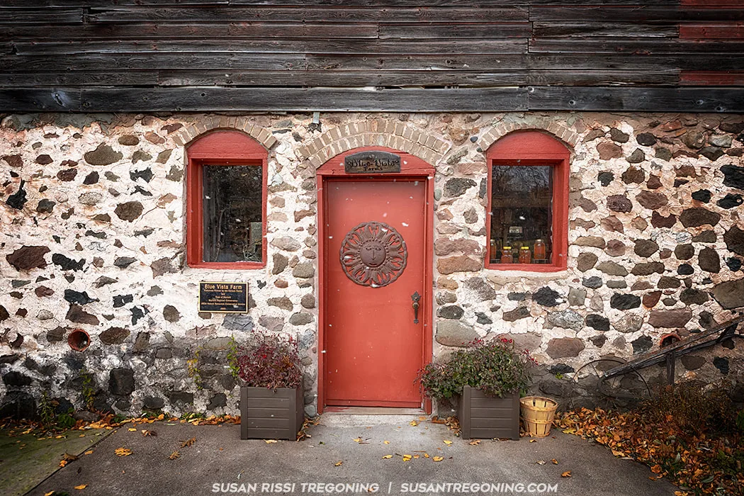 A rustic stone building with weathered wooden siding above the doorway features a red door and two red‑framed windows. The door has a decorative sun motif and an ornate handle. A small sign above the door reads “Blue Vista Farm,” and a plaque to the left also displays the farm name. Two rectangular planters flank the entrance, and a small wooden basket sits to the right. Fallen leaves are scattered on the ground, suggesting an autumn setting.