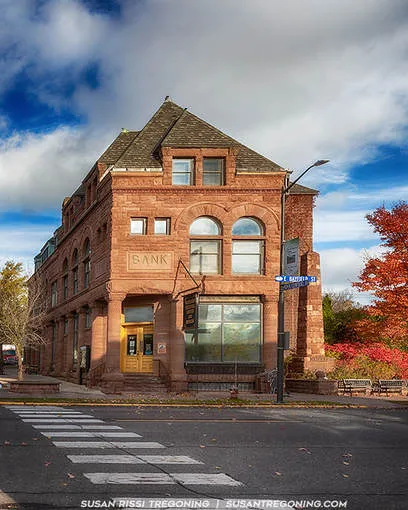   
A three‑story red sandstone building with arched second‑floor windows and a steep roof stands on a street corner. The word “BANK” is carved above the entrance. A crosswalk, lamppost, and autumn trees with red and orange leaves surround the scene under a partly cloudy sky.
