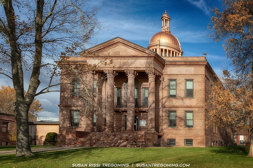 A large three‑story building made of reddish‑brown stone features a neoclassical portico with six Corinthian columns and a triangular pediment. A gold dome rises above the roof. Trees with autumn foliage and a landscaped lawn surround the structure under a partly cloudy sky.