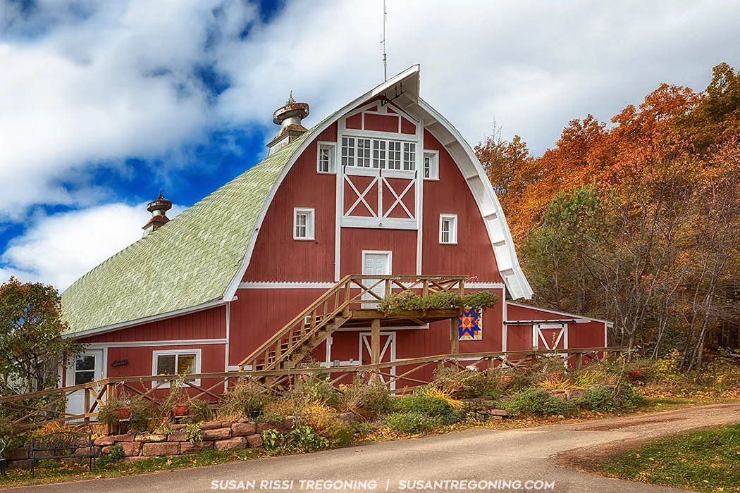A large red barn with white trim and a green gambrel roof stands beside a dirt road, surrounded by autumn trees with orange, yellow, and green leaves. The barn has multiple windows, a central upper‑level door, and a wooden staircase leading to a small balcony with a railing. A colorful quilt‑pattern decoration hangs on the front wall. The sky above is partly cloudy with patches of blue.