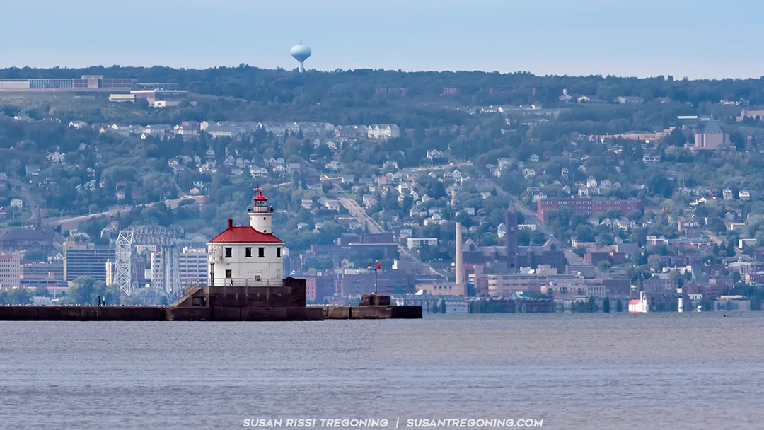 View from Wisconsin Point Beach showing the Wisconsin Point Lighthouse in the foreground, with the Superior Entry visible just beyond it. The Duluth Aerial Lift Bridge appears in the distance to the left of the Superior Entry Lighthouse, and the small silhouettes of the Duluth Ship Canal lighthouses are visible on the far right.