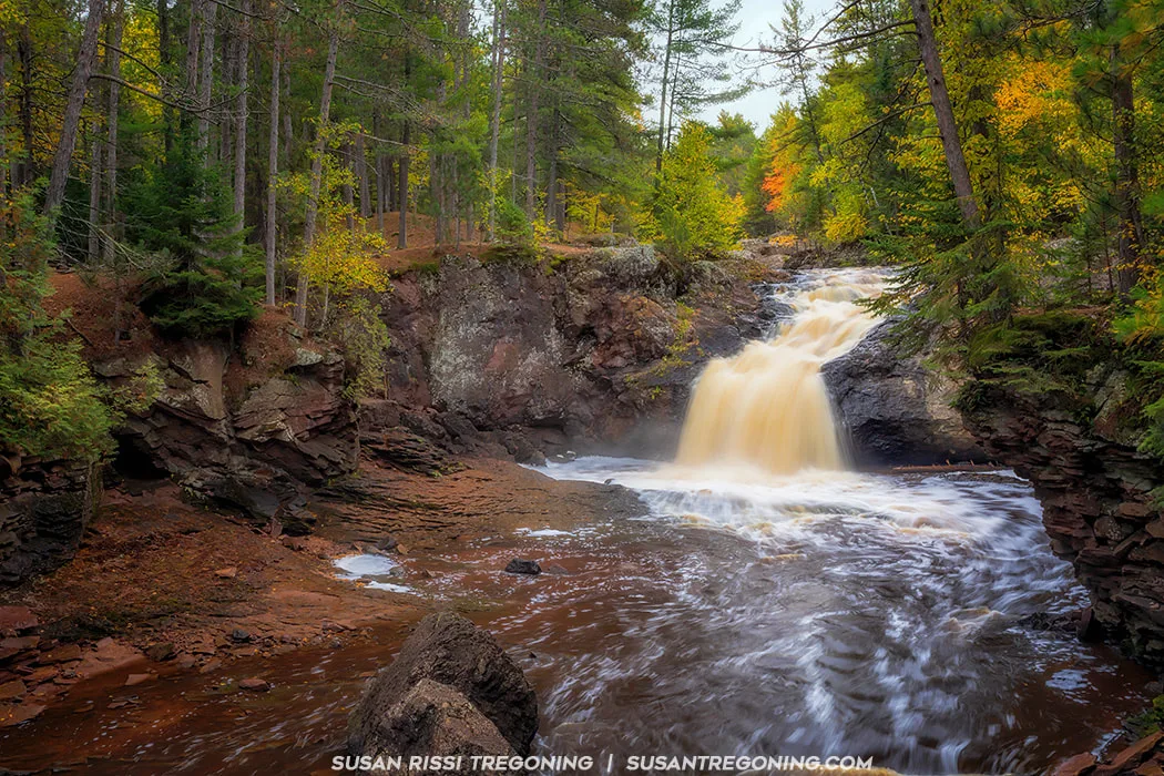 A small waterfall flows over rocky ledges into a brown, swirling pool, surrounded by a dense forest of tall trees with green and golden autumn foliage. The long-exposure effect softens the moving water, creating a smooth, misty appearance against the rugged rocks and earthy shoreline.