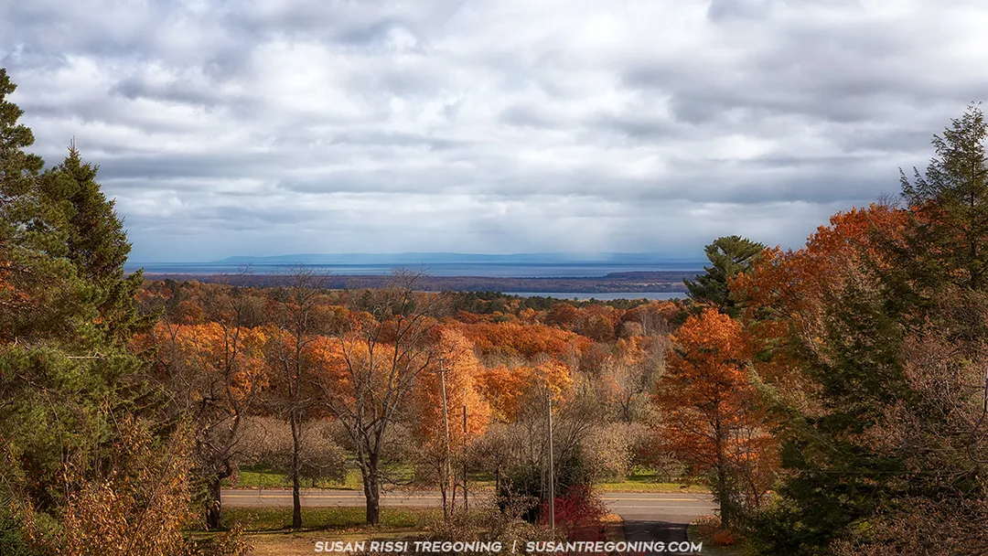 A hillside of autumn trees in shades of orange, red, yellow, and green stretches toward a large body of water in the distance. A paved road runs across the foreground, and the sky above is cloudy with soft light filtering through.