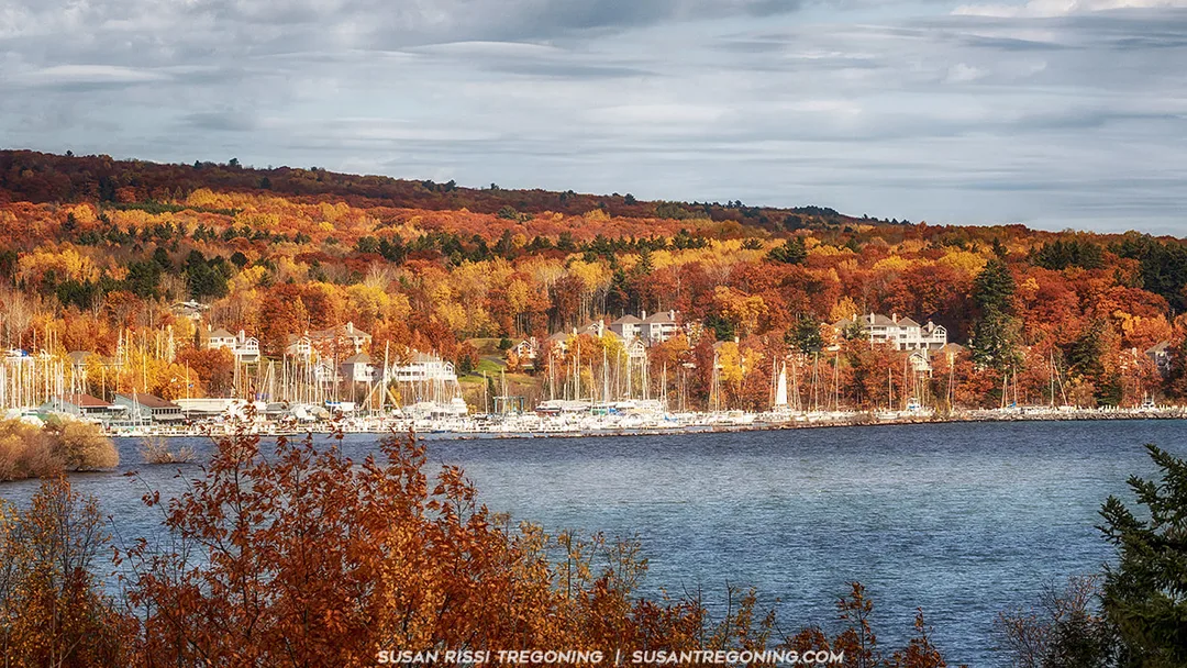A lakeside marina sits along the shoreline with numerous sailboats docked in calm water. Behind the marina, houses and trees with vibrant red, orange, and yellow autumn foliage rise up a forested hillside. A partly cloudy sky stretches above the scene.
