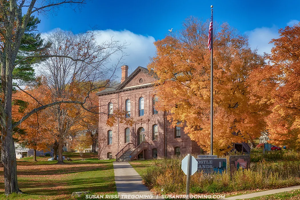   
A historic brownstone building with a gabled roof and arched windows stands surrounded by bright autumn trees in shades of orange and yellow. A central staircase leads to the main entrance, and an American flag flies on a flagpole in front. A sign near the sidewalk reads “Visitor Center.” The sky is partly cloudy.