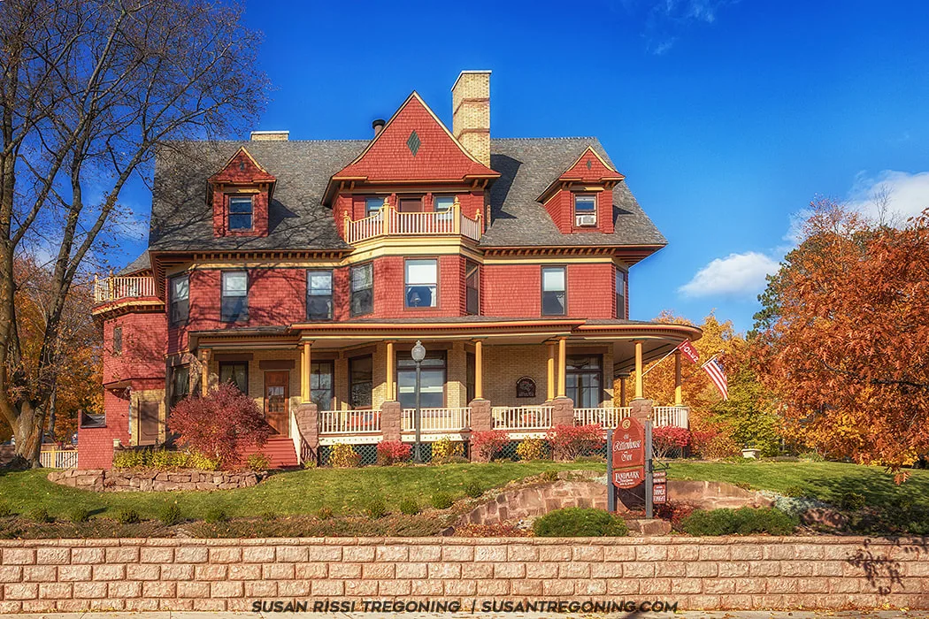 A large Victorian house with red and tan exterior walls, steep gabled roofs, and multiple dormers stands behind a stone retaining wall. A wraparound porch with yellow columns and decorative railings extends across the front. A sign in the yard reads “Redstone Inn” with “Luxury Inn” below it. An American flag hangs from the porch, and autumn trees with orange and yellow leaves surround the house under a bright blue sky.