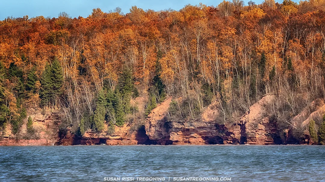 A rocky shoreline with reddish‑brown cliffs rises above calm blue water. The forest above the cliffs is filled with autumn foliage in shades of orange, yellow, and brown, with a few evergreen trees mixed in. The colorful trees cover the hillside, creating a vibrant fall landscape against the lake.