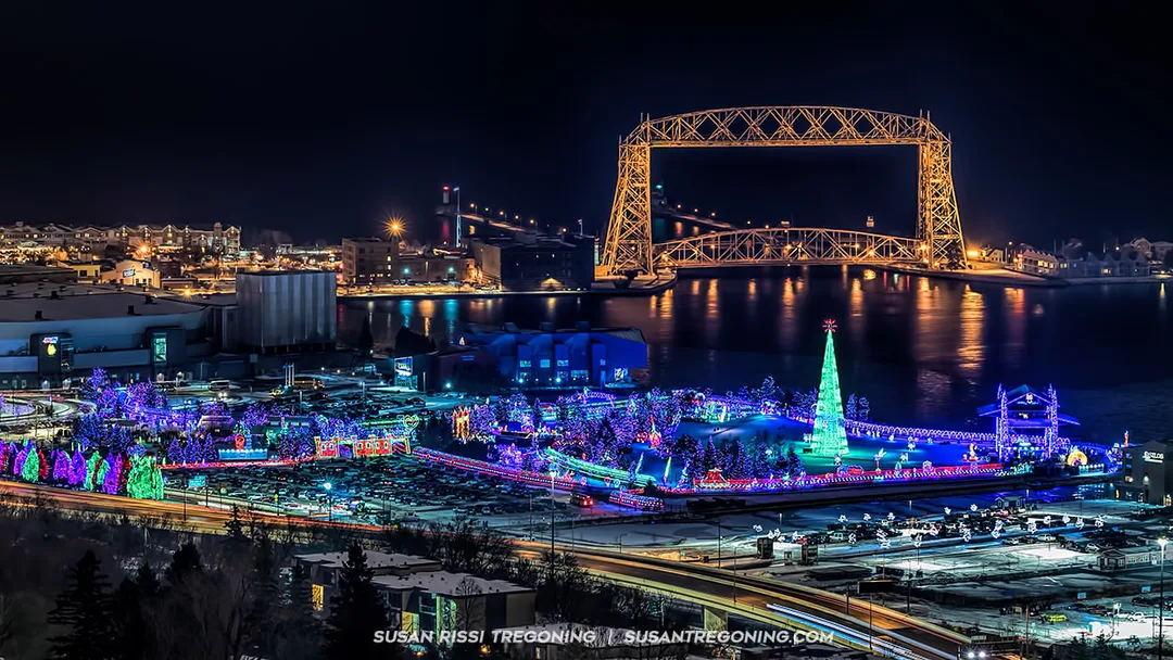 A nighttime view of a brightly lit holiday display with colorful trees, decorations, and a tall cone‑shaped Christmas tree structure in the foreground. Behind the display, the Duluth Aerial Lift Bridge is illuminated and reflected on the water, with additional city lights visible across the shoreline.