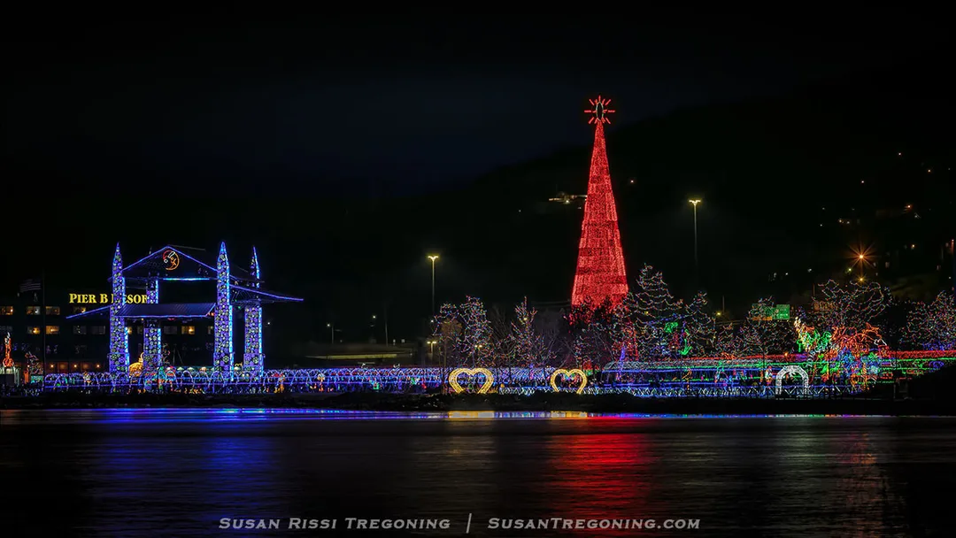 A nighttime waterfront scene featuring colorful holiday light displays reflected on the water. On the left, a structure outlined in blue and white lights reads “PIER B.” To the right, a tall red cone‑shaped Christmas tree made of lights stands topped with a star, surrounded by additional illuminated decorations including heart shapes. Buildings and other lit elements frame the festive area along the shoreline.