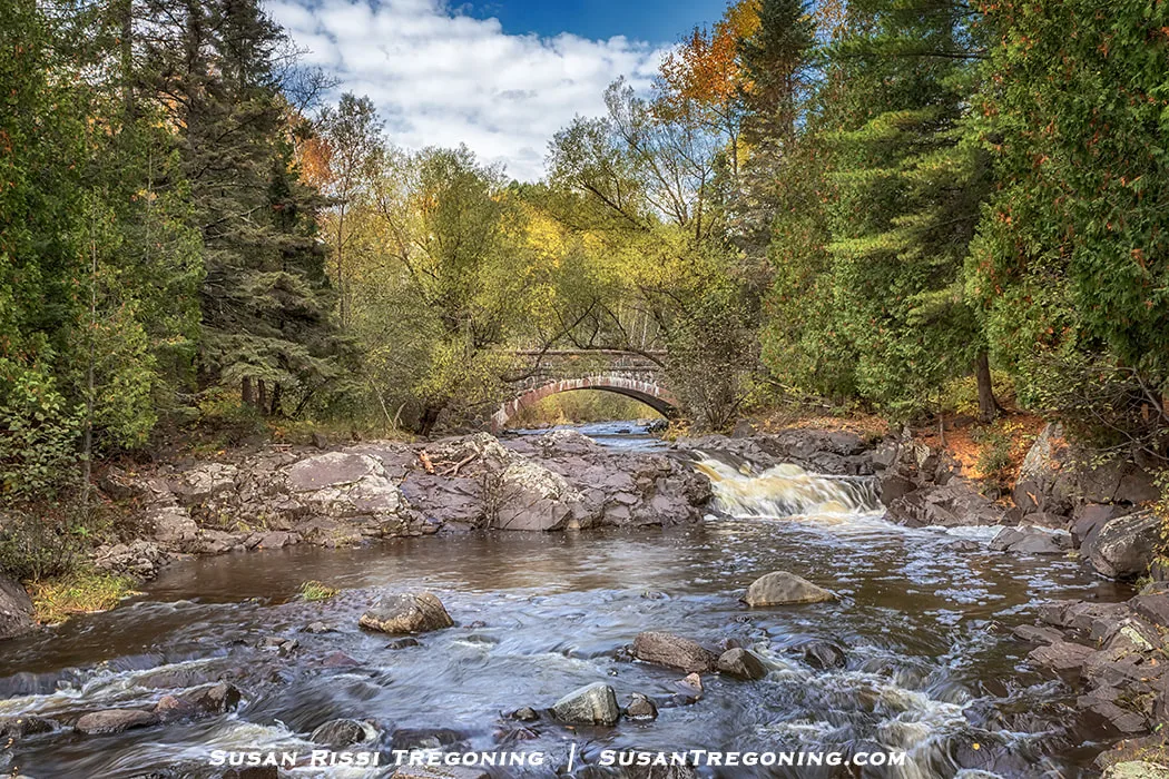Bridge #1 on Seven Bridges Road arches over Amity Creek in Duluth, Minnesota. The stone bridge rises above rushing water and dark rocks, surrounded by dense green and early autumn foliage beneath a partly cloudy sky.