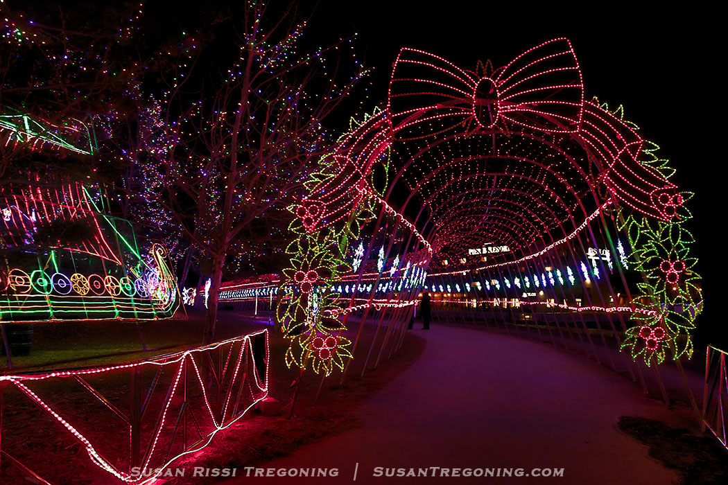 A nighttime holiday light display featuring a large illuminated archway decorated with red and green lights forming a bow and floral patterns over a pathway. To the left, a multicolored light sculpture resembling a train or sleigh sits near the walkway. Trees along the path are wrapped in small white lights, with additional festive decorations visible in the background.