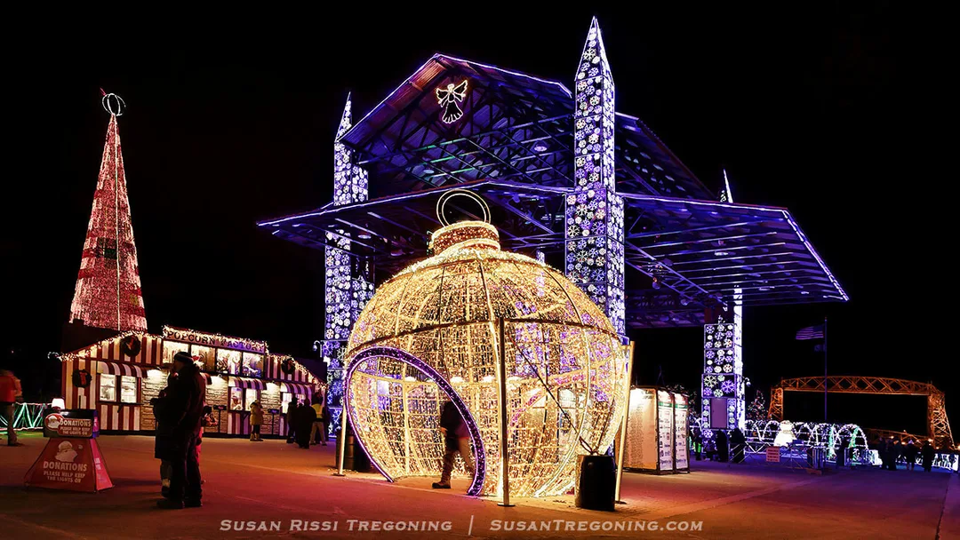 A glowing ornament tunnel and nearby lighted displays brighten the holiday walkway, with a purple‑lit pavilion and festive tree adding color to the night.
