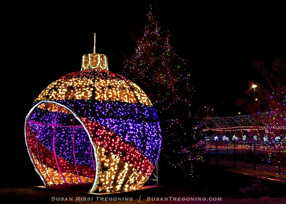 A large illuminated Christmas ornament made of gold, red, and blue lights stands open for visitors to walk through. Behind it, a tall Christmas tree decorated with multicolored lights rises above the display. Additional festive light installations fill the background, including a canopy of lights and other holiday decorations, all set against the night sky.