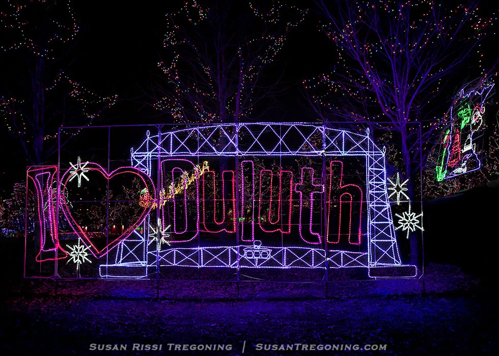   
A nighttime holiday light display featuring the phrase “I ♥ Duluth” formed with colorful lights. The design includes a red and white heart symbol and an outline of the Aerial Lift Bridge in blue lights around the word “Duluth.” Snowflake shapes and trees decorated with multicolored lights appear in the background.