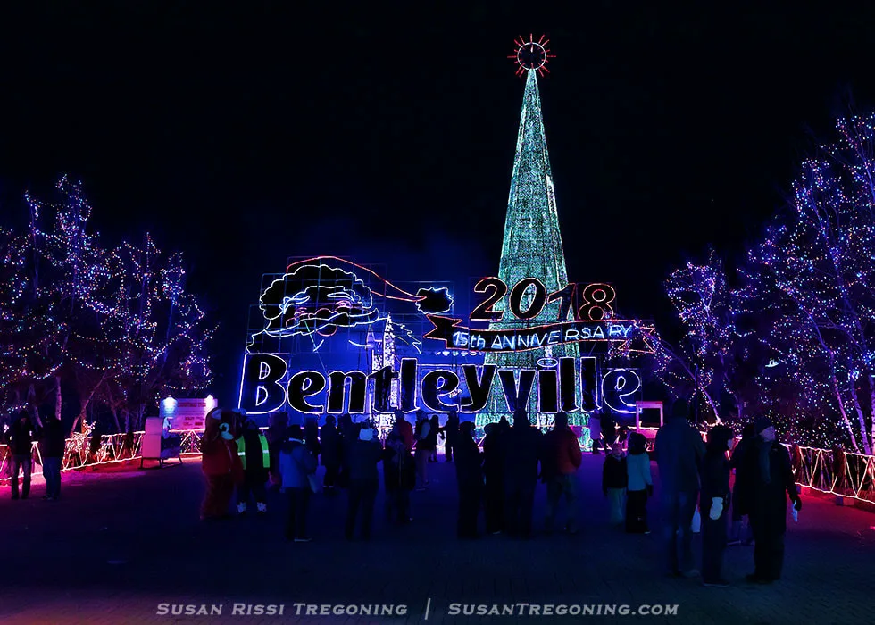 Nighttime holiday light display at Bentleyville featuring a tall cone‑shaped Christmas tree made of lights topped with a red star. In front of the tree, an illuminated sign reads “2018,” “15th Anniversary,” and “Bentleyville,” with Santa’s face outlined in lights above the text. Colorful lights decorate nearby trees, and a crowd of visitors stands in the foreground viewing and photographing the display.