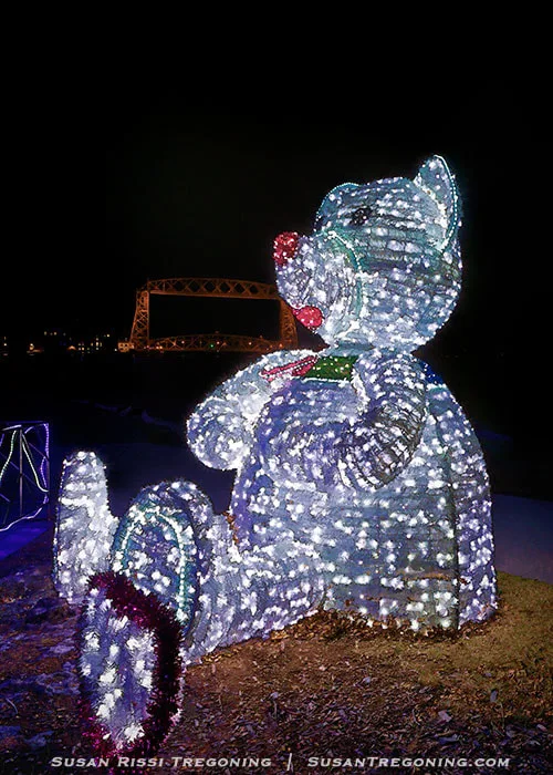 A large bear sculpture made of white lights sits upright outdoors at night, with a red‑lit nose and green lights on its chest. Behind the bear, a bridge outlined in lights spans the background, and the surrounding area is dim except for the illuminated display.