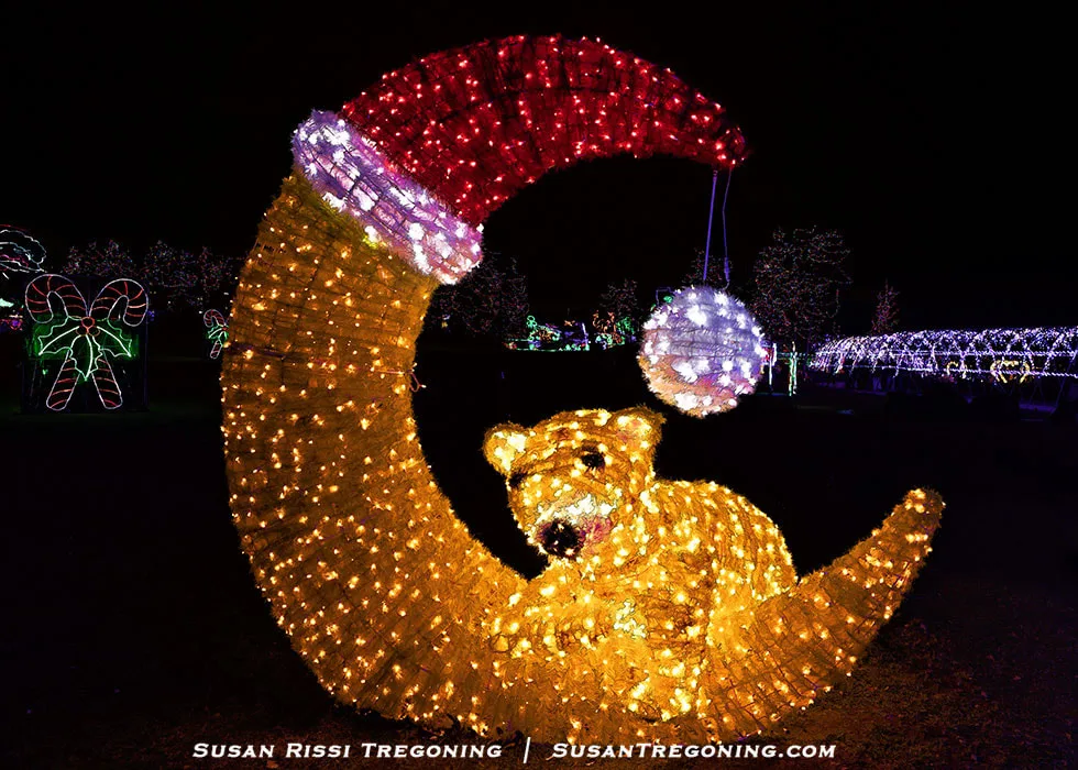 A large illuminated sculpture of a bear lying on a crescent moon glows against the night. Both the bear and moon are made of yellow lights, and the moon is topped with a red and white Santa hat. Additional holiday light displays, including candy canes and tunnel‑like arches, appear in the background.