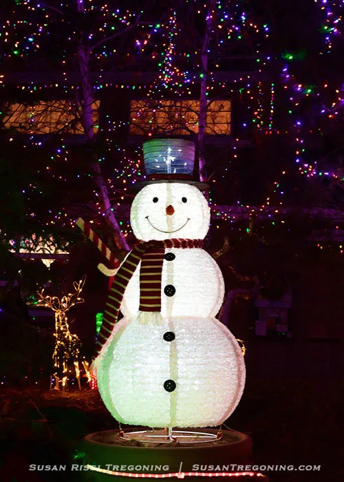 A brightly lit snowman decoration stands outdoors at night, with a glowing top hat, striped scarf, and illuminated buttons. A lighted reindeer figure appears to the left, and trees and a nearby building are covered in colorful holiday lights in the background.