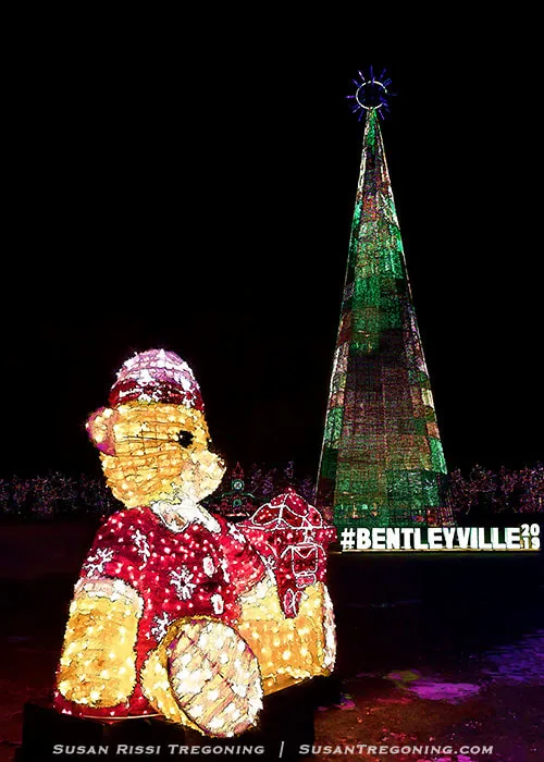 A large illuminated teddy bear wearing a red outfit and hat holds a wrapped gift in the foreground of a nighttime holiday display. Behind it, a tall cone‑shaped Christmas tree made of green and multicolored lights rises into the sky, topped with a blue star. The illuminated text “#BENTLEYVILLE 2019” appears near the base of the tree, with additional festive lights visible in the background.